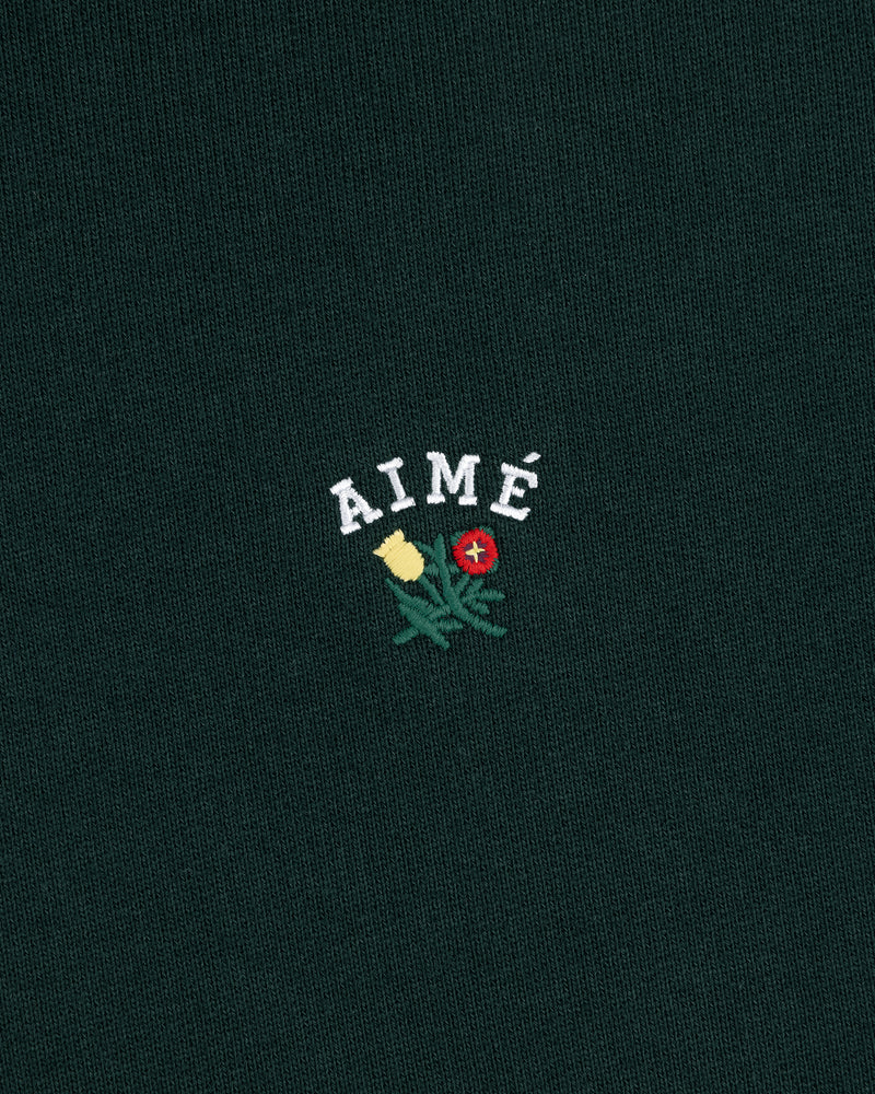 The Crest Hoodie features "AIMÉ" embroidered above yellow and red flowers on dark green fabric.