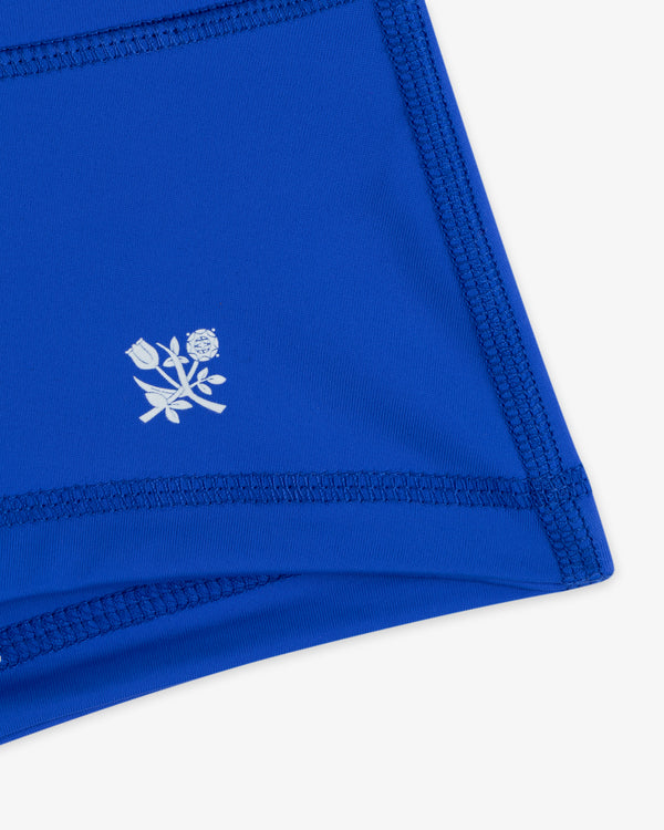 Close-up of Crest Swim Short in blue, featuring dark stitching and a small white floral emblem.