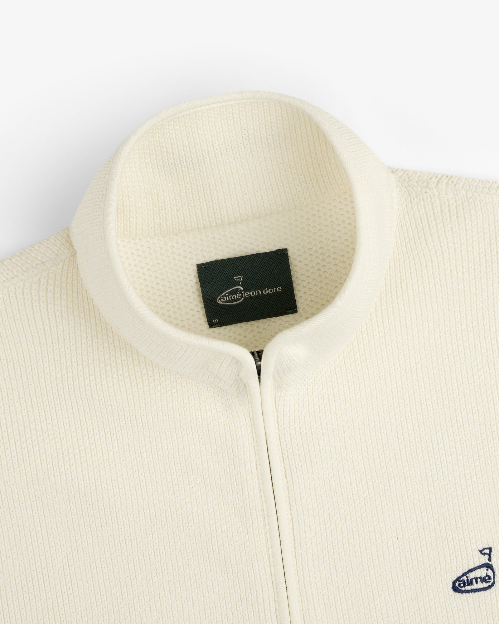 Close-up of the ALD Golf Piqué Full Zip Sweater with a ribbed texture and small navy Aimé Leon Dore chest logo.