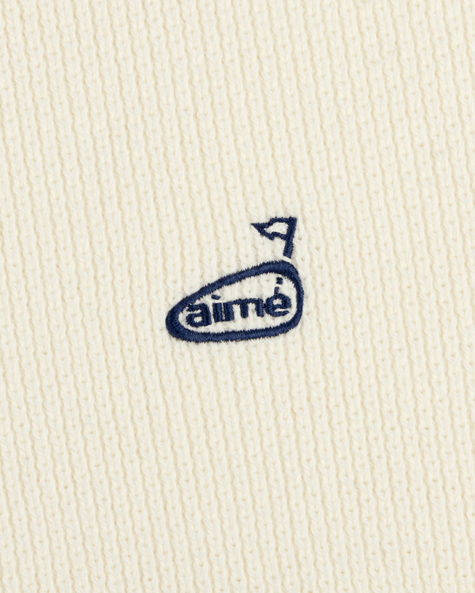 Close-up of the ALD Golf Piqué Full Zip Sweater with a cream knit and small blue embroidered aimé logo with an arrow.
