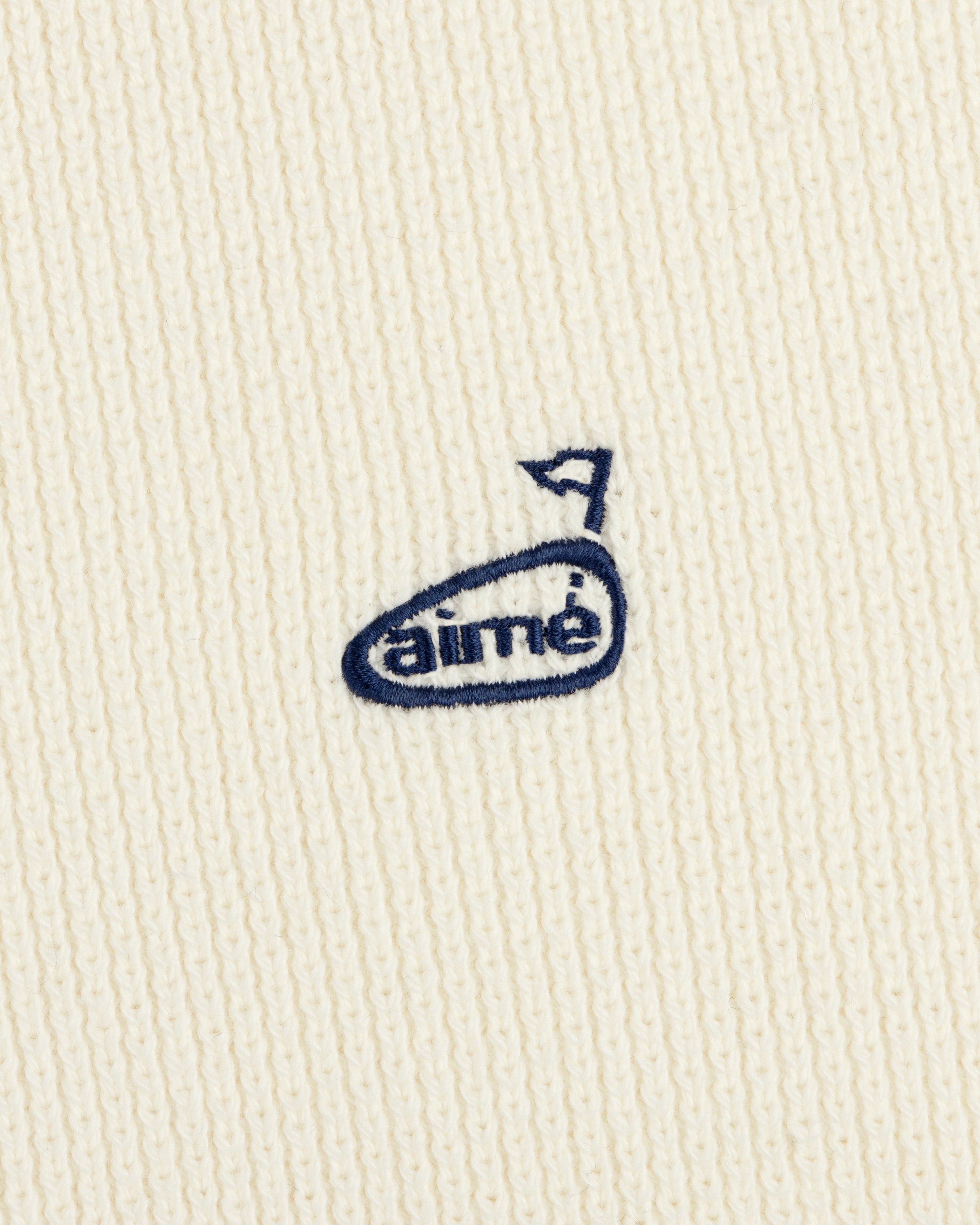 Close-up of the ALD Golf Piqué Full Zip Sweater with a cream knit and small blue embroidered aimé logo with an arrow.