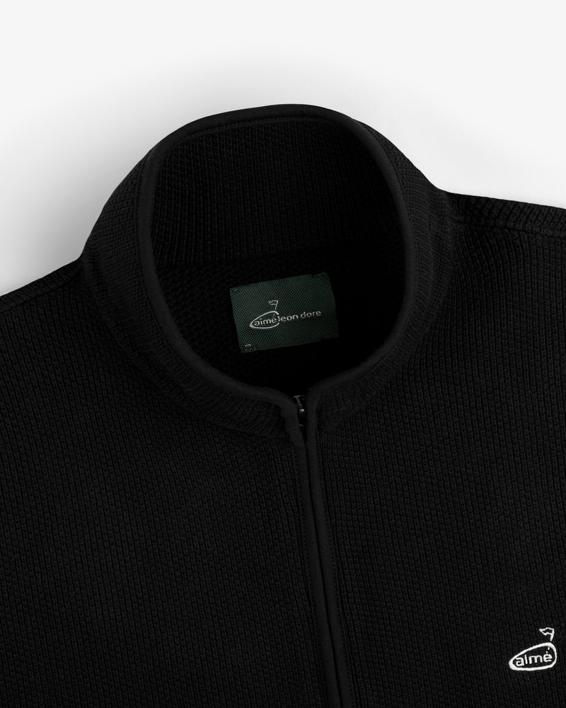 Close-up of the ALD Golf Piqué Full Zip Sweater, black with ribbed texture and Aimé Leon Dore logo on the chest.