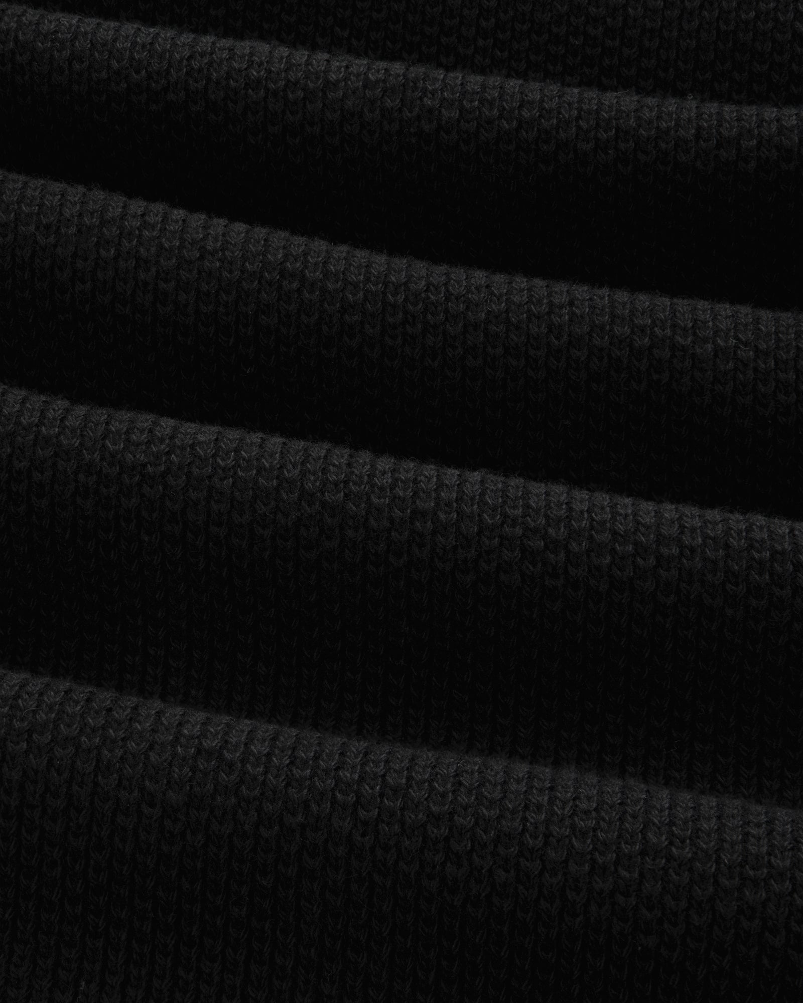 Close-up of ALD Golf Piqué Full Zip Sweater in black, showing ribbed fabric with horizontal folds and textured knit.