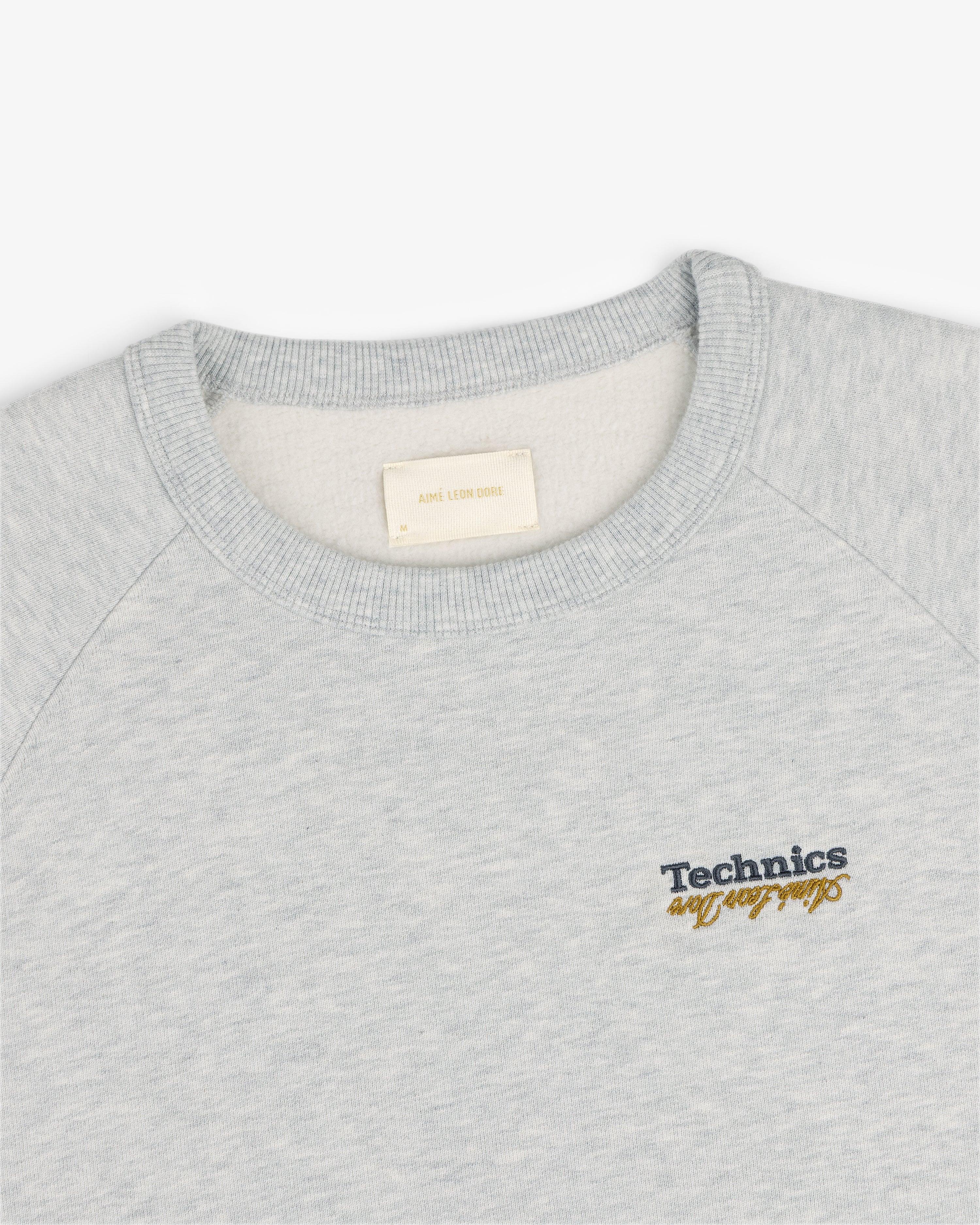ALD / Technics World Champions Crewneck in light gray with black and yellow embroidered Technics cursive chest text.