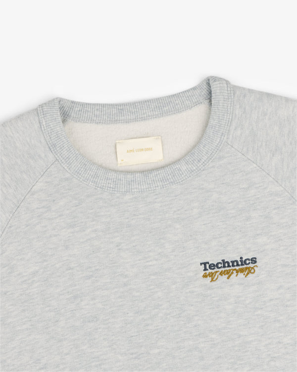 ALD / Technics World Champions Crewneck in light gray with black and yellow embroidered Technics cursive chest text.