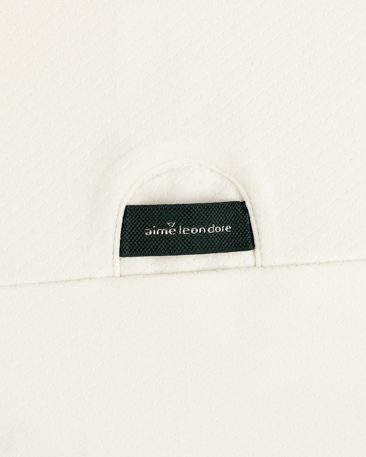 A close-up of a black Aimé Leon Dore label sewn onto the off-white ALD Golf Quarter Zip Pullover’s textured fabric.