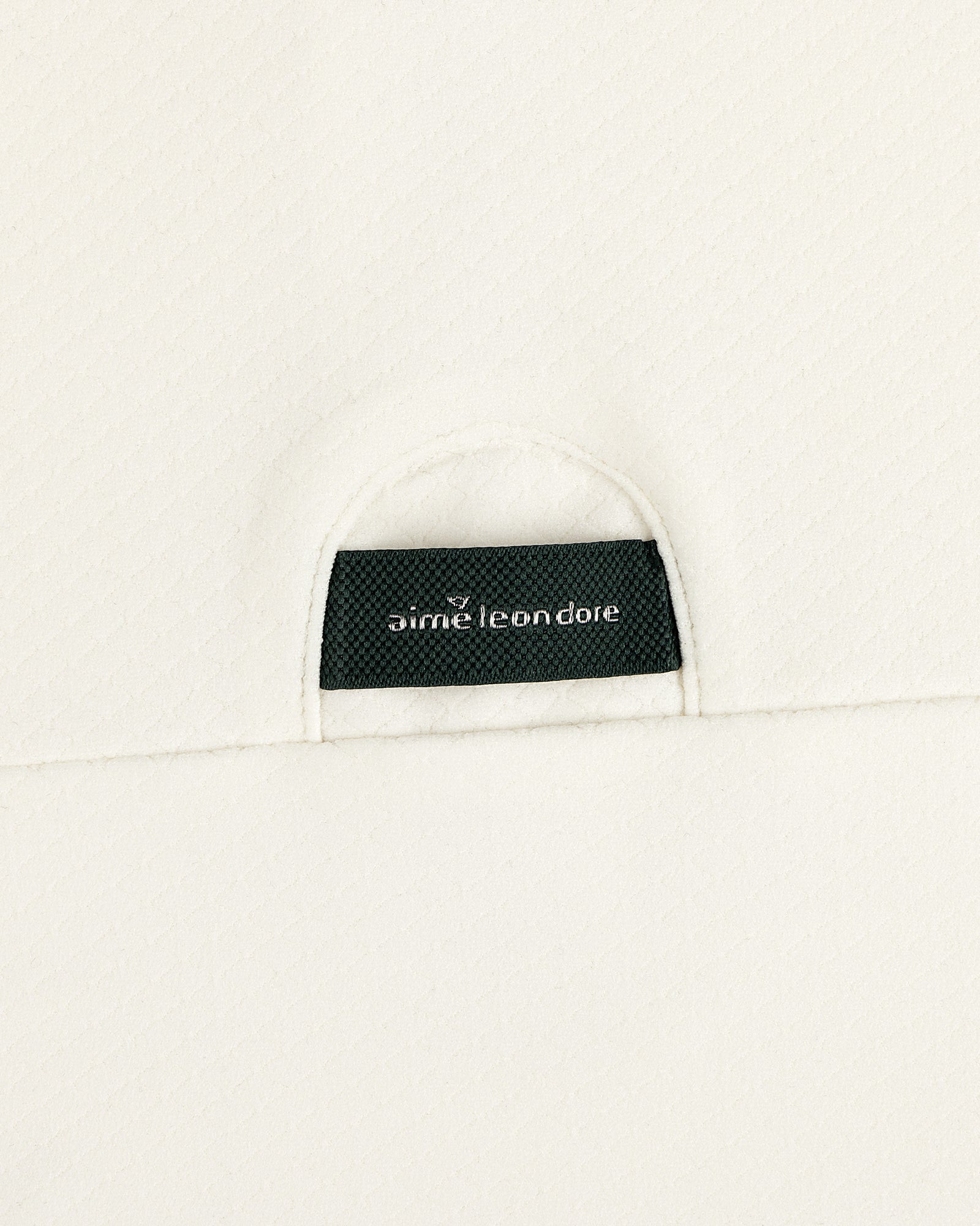 A close-up of a black Aimé Leon Dore label sewn onto the off-white ALD Golf Quarter Zip Pullover’s textured fabric.