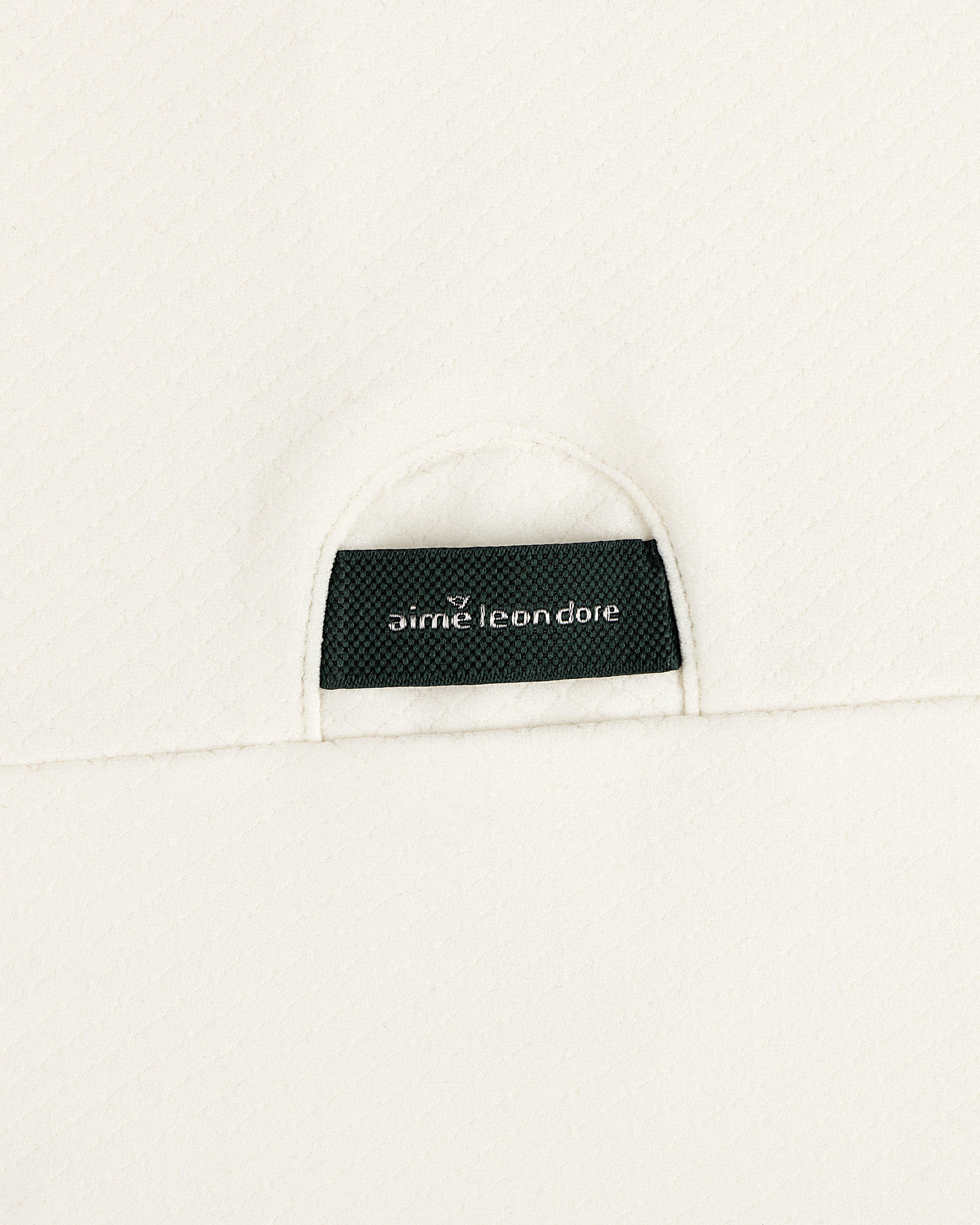 A close-up of a black Aimé Leon Dore label sewn onto the off-white ALD Golf Quarter Zip Pullover’s textured fabric.