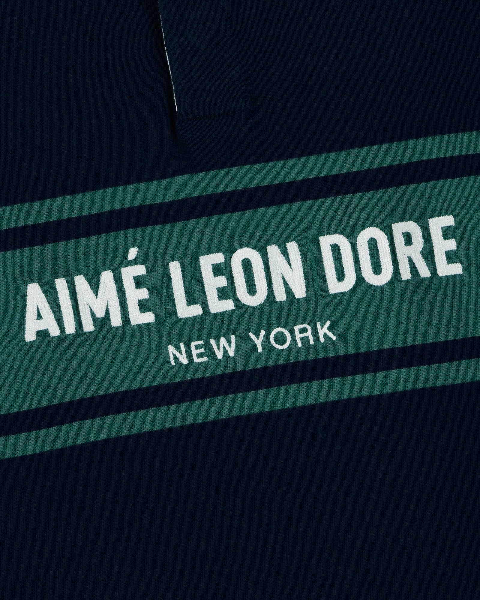 Close-up of the Chest Stripe Logo Rugby with AIMÉ LEON DORE NEW YORK printed on green and navy blue fabric.