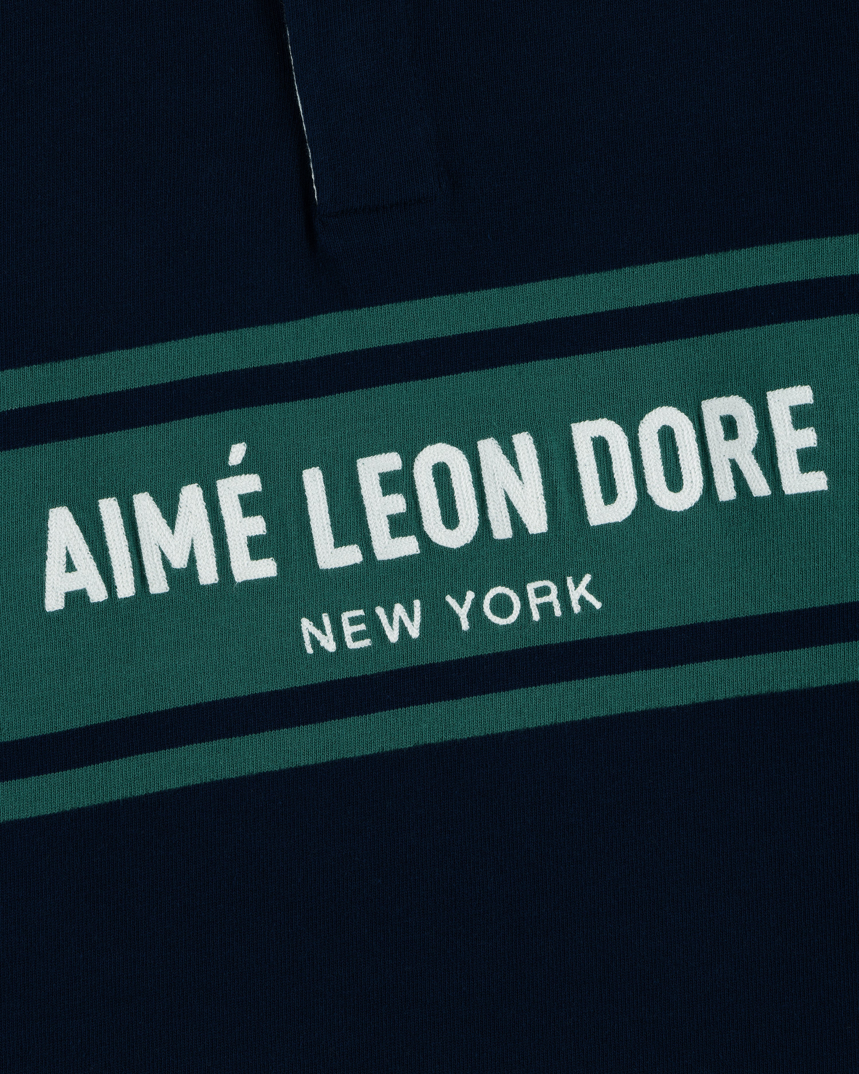 Close-up of the Chest Stripe Logo Rugby with AIMÉ LEON DORE NEW YORK printed on green and navy blue fabric.