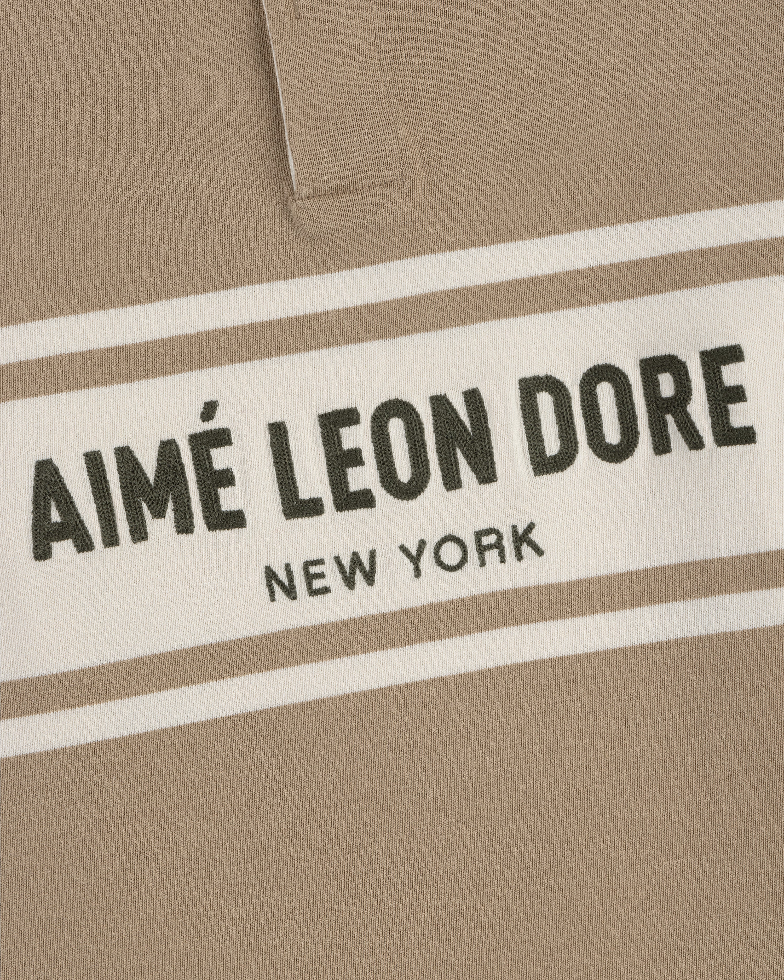 Close-up of the Chest Stripe Logo Rugby, a beige shirt with bold AIMÉ LEON DORE NEW YORK text on a white chest stripe.