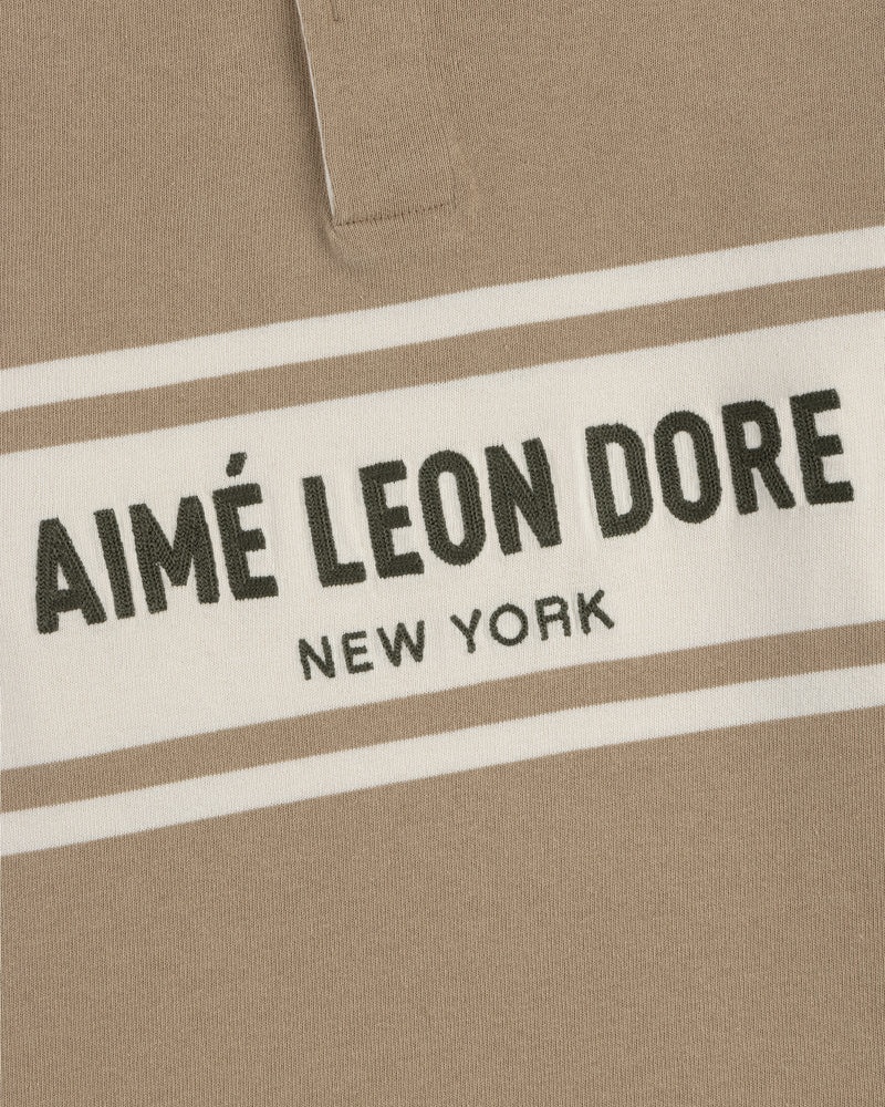 Close-up of the Chest Stripe Logo Rugby, a beige shirt with bold AIMÉ LEON DORE NEW YORK text on a white chest stripe.