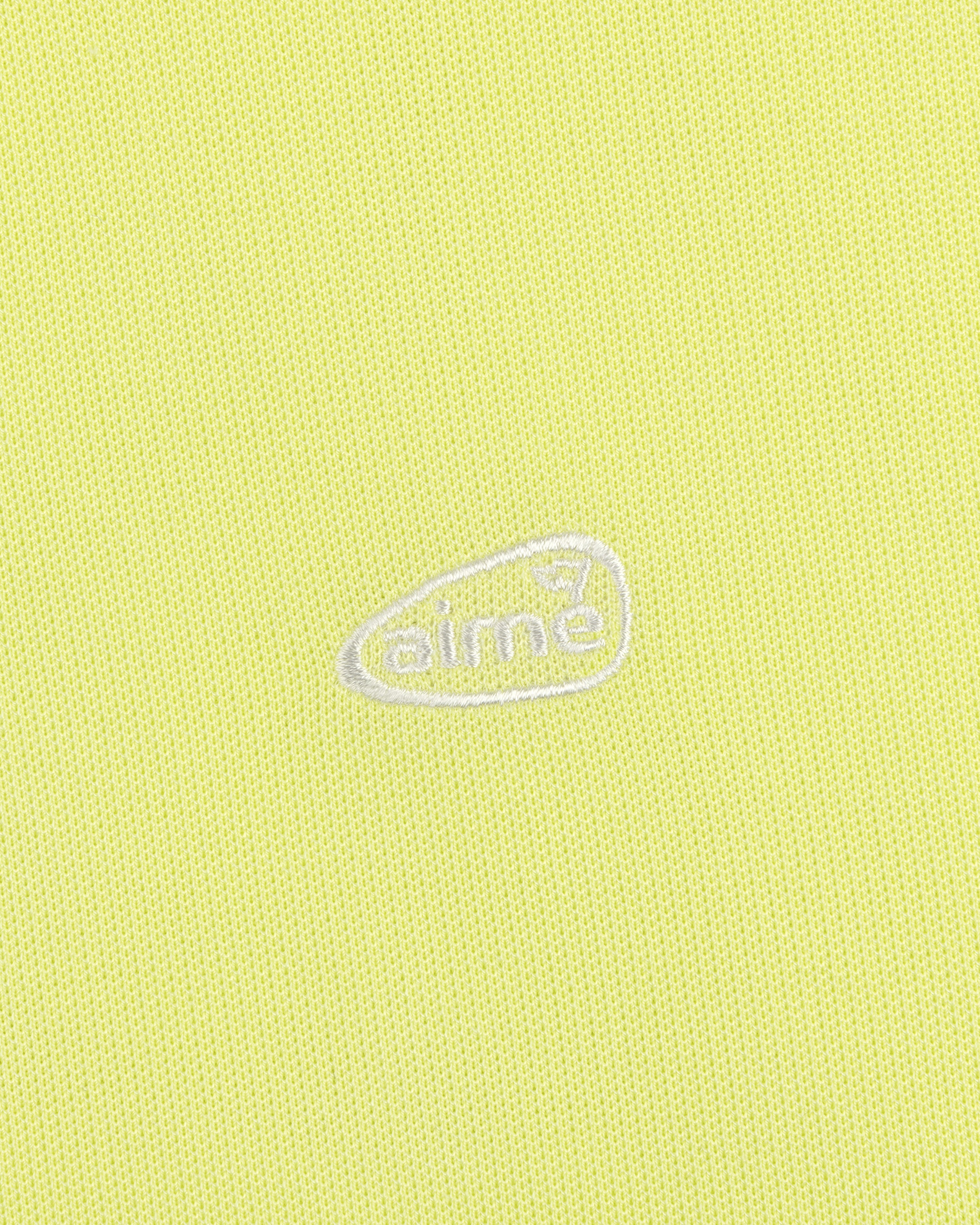 Yellow textured ALD Golf Polo with small white aimé logo inside an irregular oval at the center.