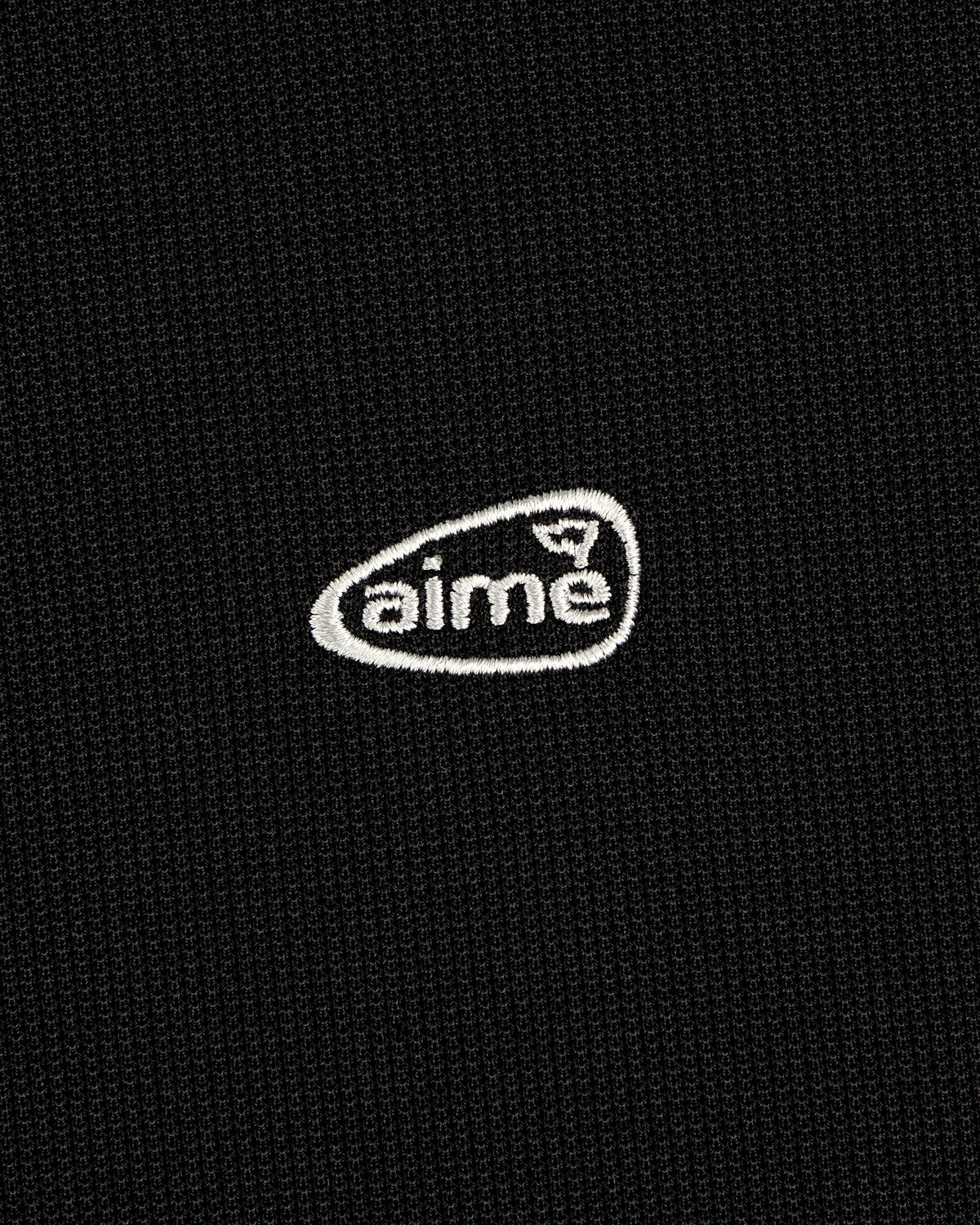 A small white aime logo is embroidered on a textured black ALD Golf Polo.