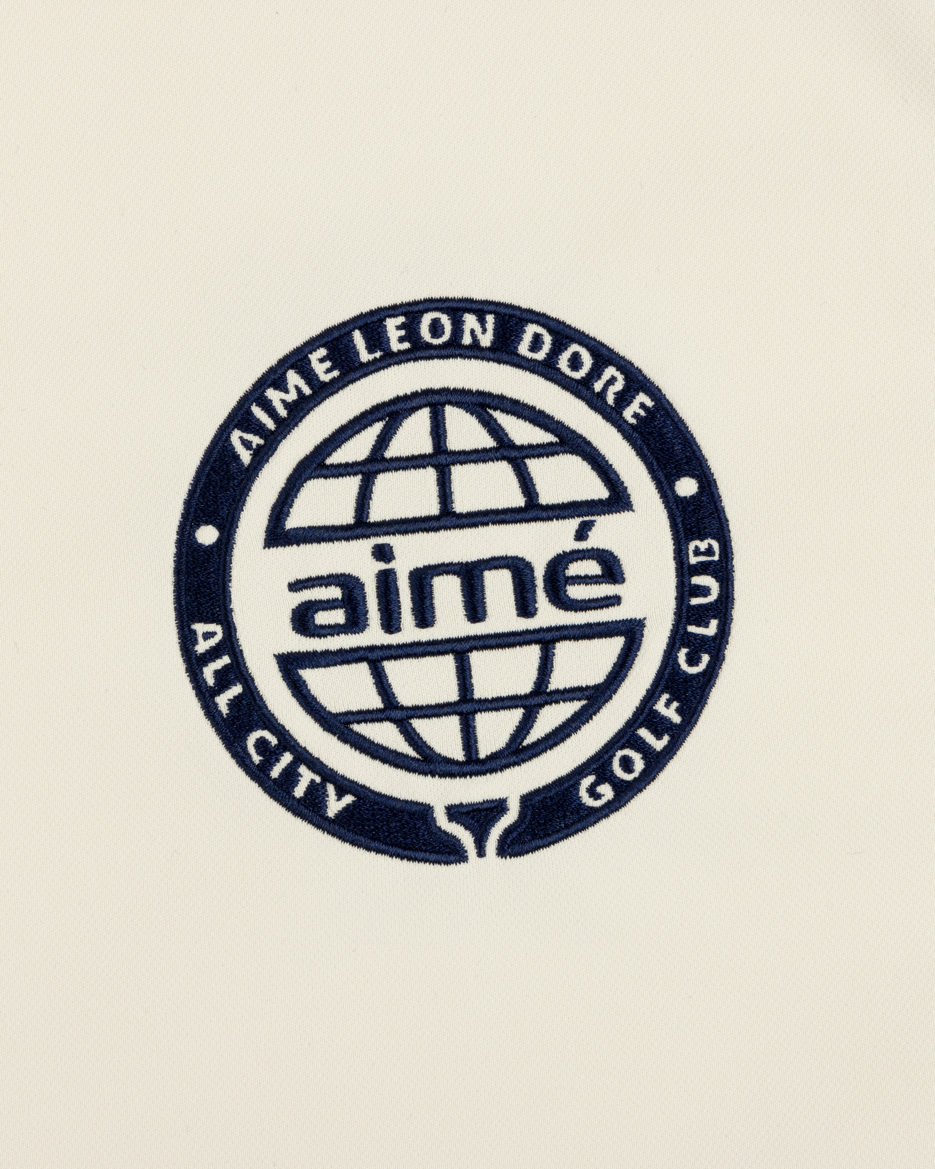 Navy blue globe logo with “AIMÉ LEON DORE ALL CITY GOLF CLUB” and “aimé” on cream ALD Golf Tour Polo.