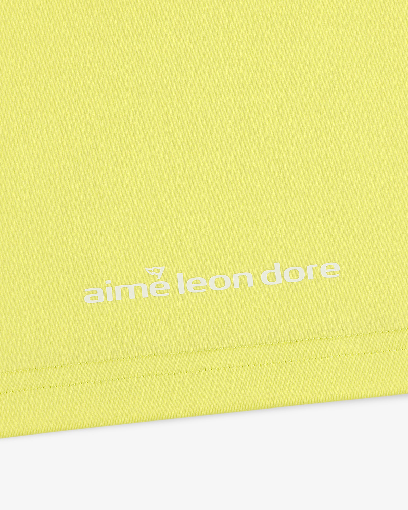 Close-up of a yellow ALD Golf Performance Tee with "aime leon dore" printed in white near the bottom edge.