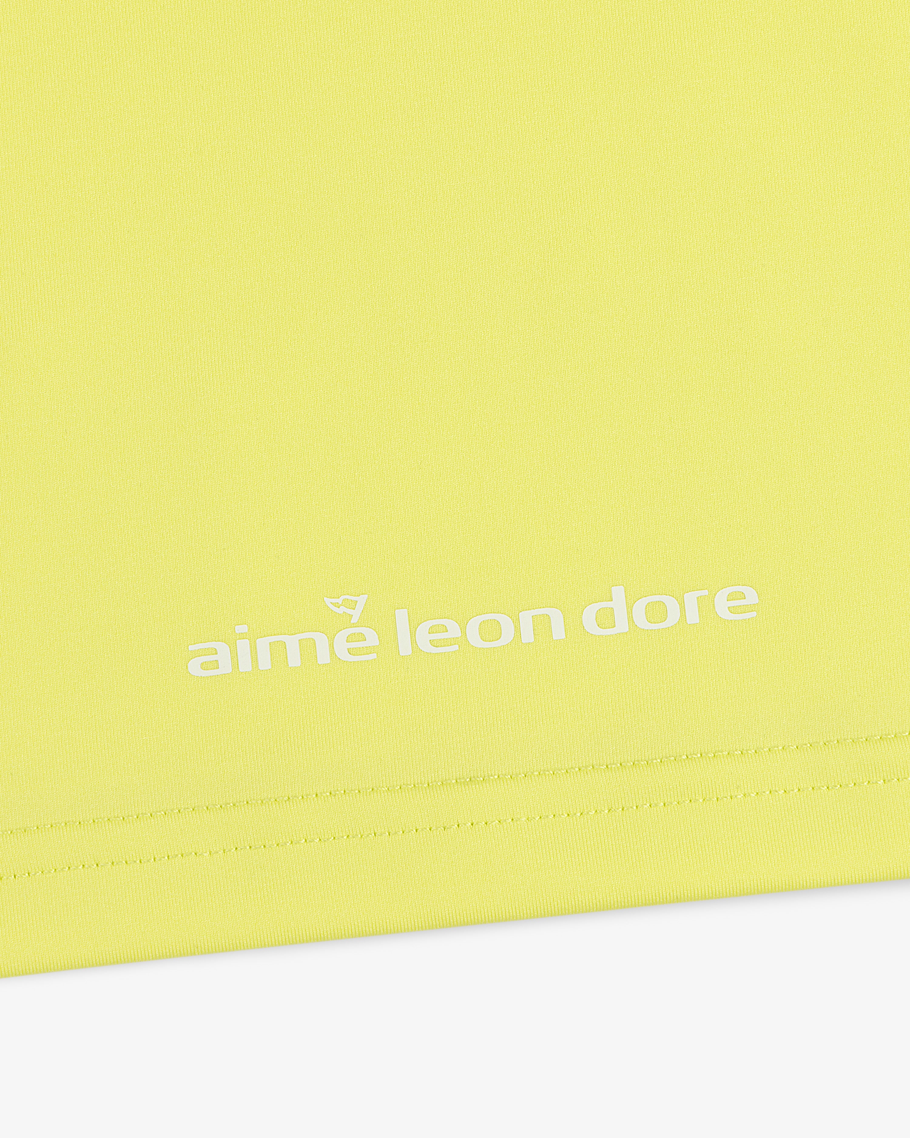 Close-up of a yellow ALD Golf Performance Tee with "aime leon dore" printed in white near the bottom edge.