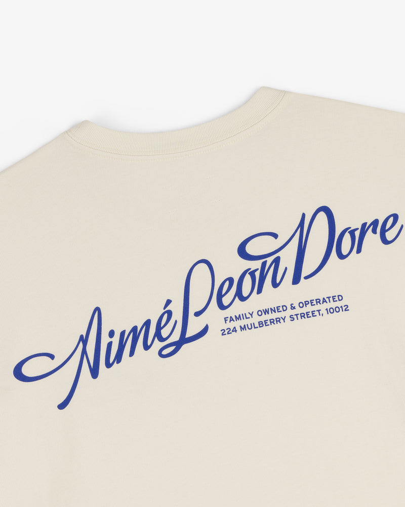 Beige Pappoús Logo Tee with large blue Aimé Leon Dore script and 224 Mulberry Street, 10012 in small text.