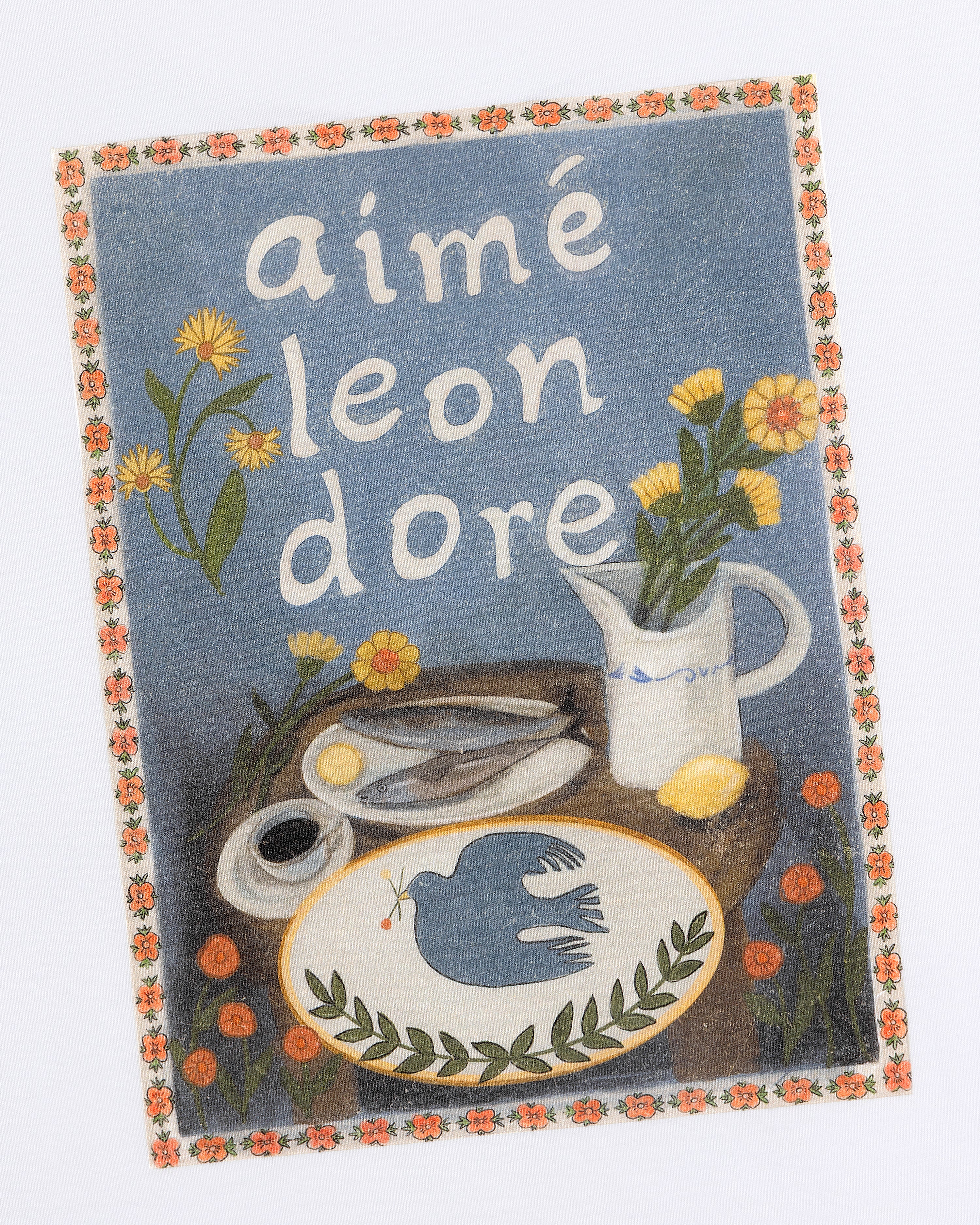 The Breakfast Tee features a vintage-style print with aimé leon dore, flowers, scissors, and a blue bird on a table.
