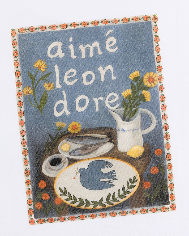 The Breakfast Tee features a vintage-style print with aimé leon dore, flowers, scissors, and a blue bird on a table.