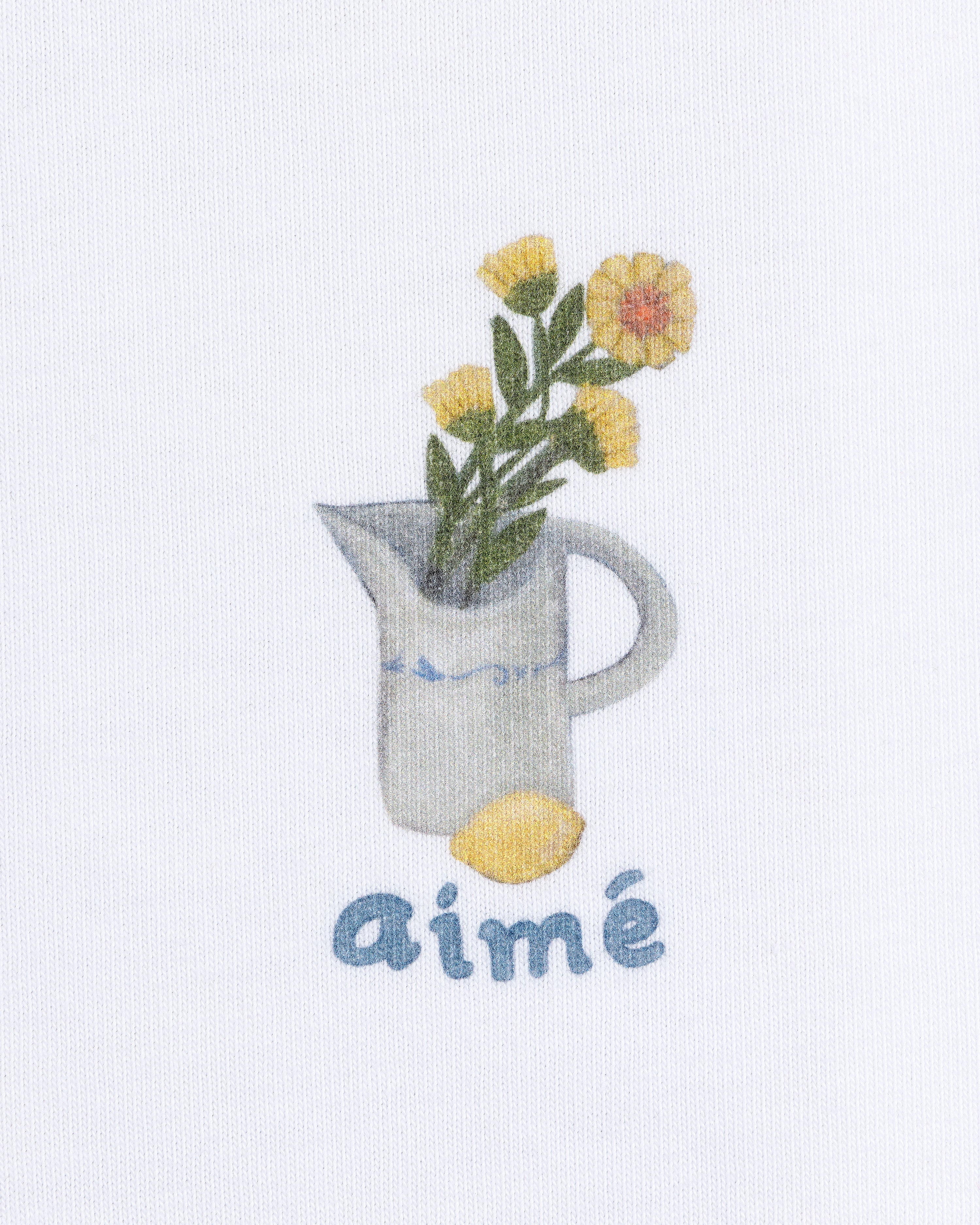 Breakfast Tee featuring a yellow flower pitcher and lemon with "aimé" below on a white background.