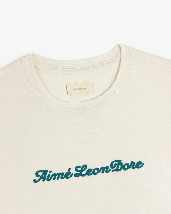 Cream-colored Puff Print Slub Logo Tee with Aimé Leon Dore in teal script across the chest.