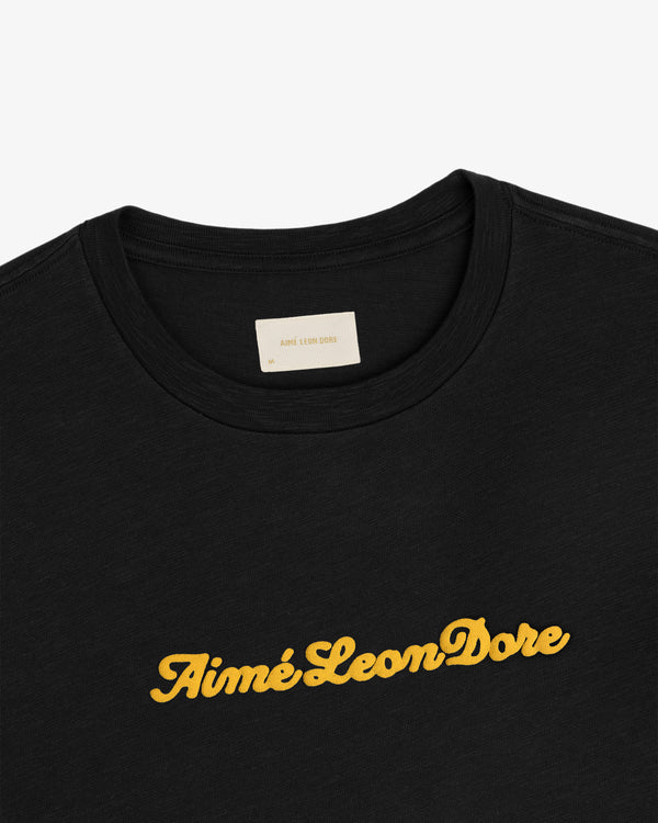 Puff Print Slub Logo Tee: black t-shirt with Aimé Leon Dore in yellow script across the chest.