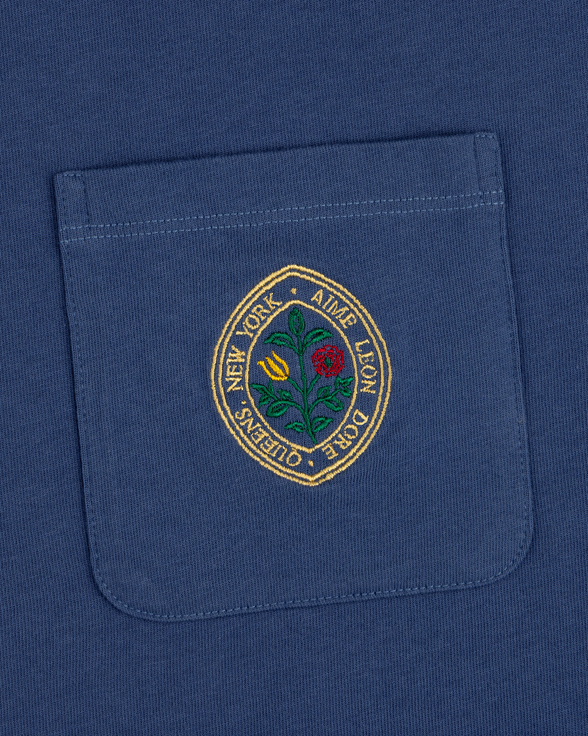 The Botanical Crest Pocket Tee features a blue pocket with a round embroidered yellow and red flower patch and border text.