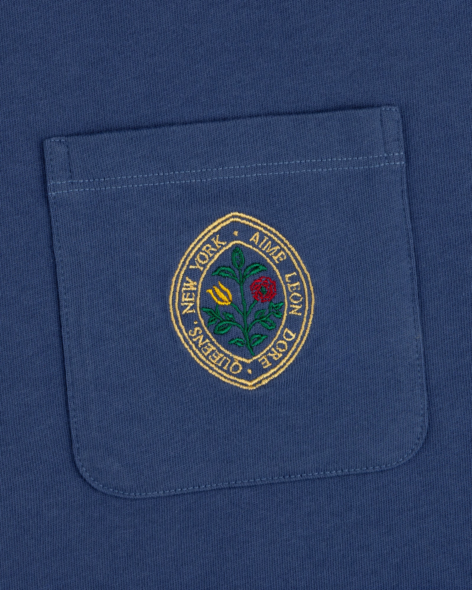 The Botanical Crest Pocket Tee features a blue pocket with a round embroidered yellow and red flower patch and border text.