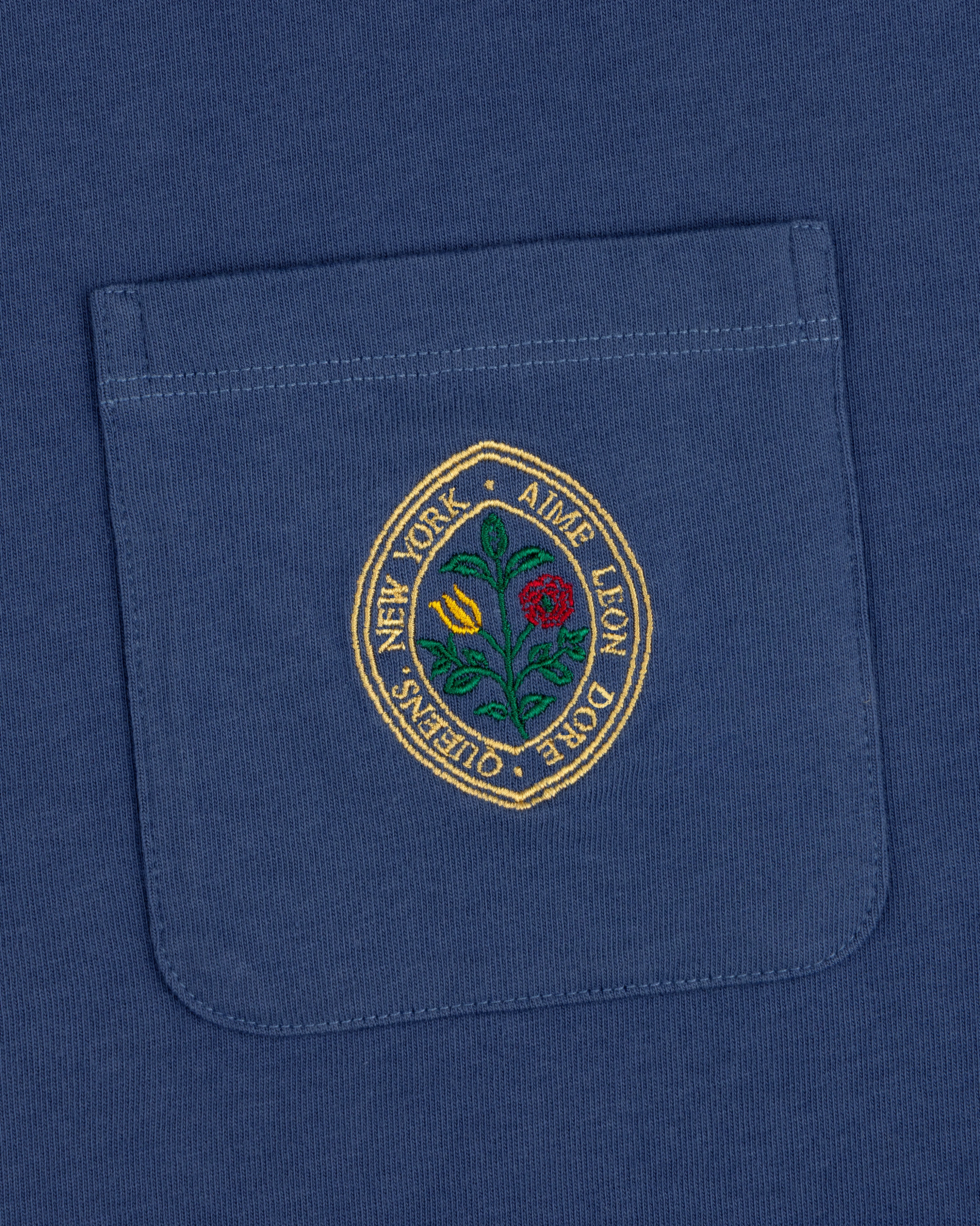 The Botanical Crest Pocket Tee features a blue pocket with a round embroidered yellow and red flower patch and border text.