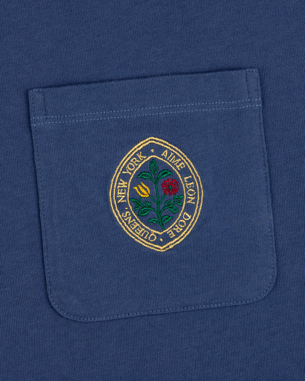 The Botanical Crest Pocket Tee features a blue pocket with a round embroidered yellow and red flower patch and border text.