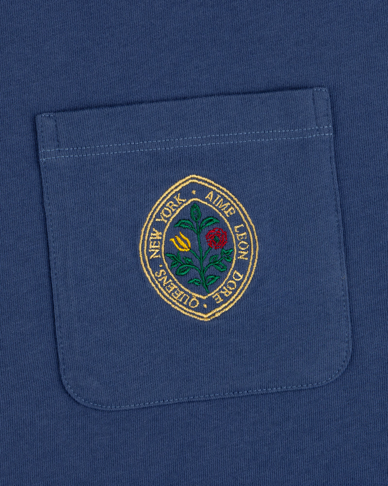 The Botanical Crest Pocket Tee features a blue pocket with a round embroidered yellow and red flower patch and border text.