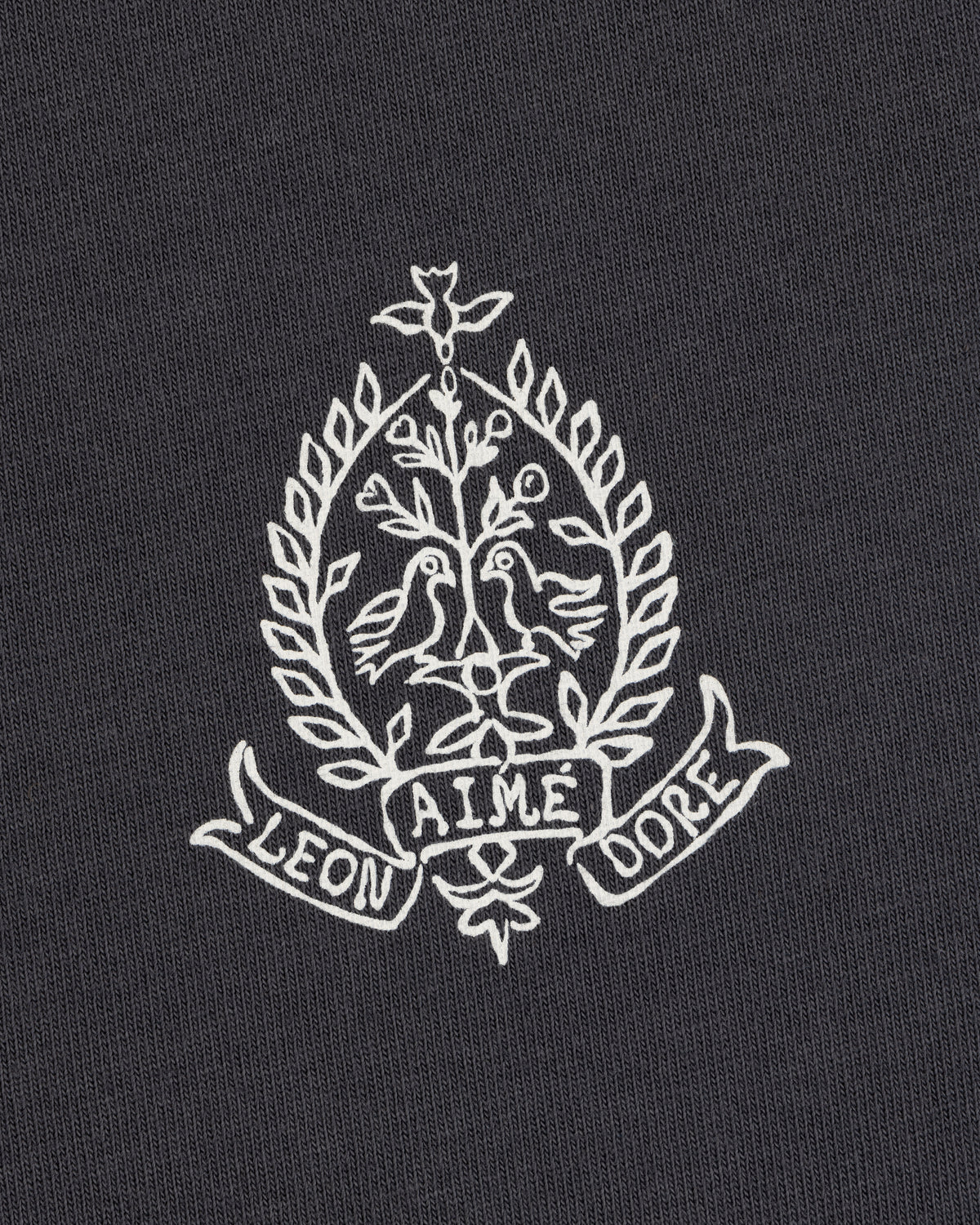 White crest with two birds, leaves, and "LEON AIME DOPE" on dark Saint George Graphic Tee.