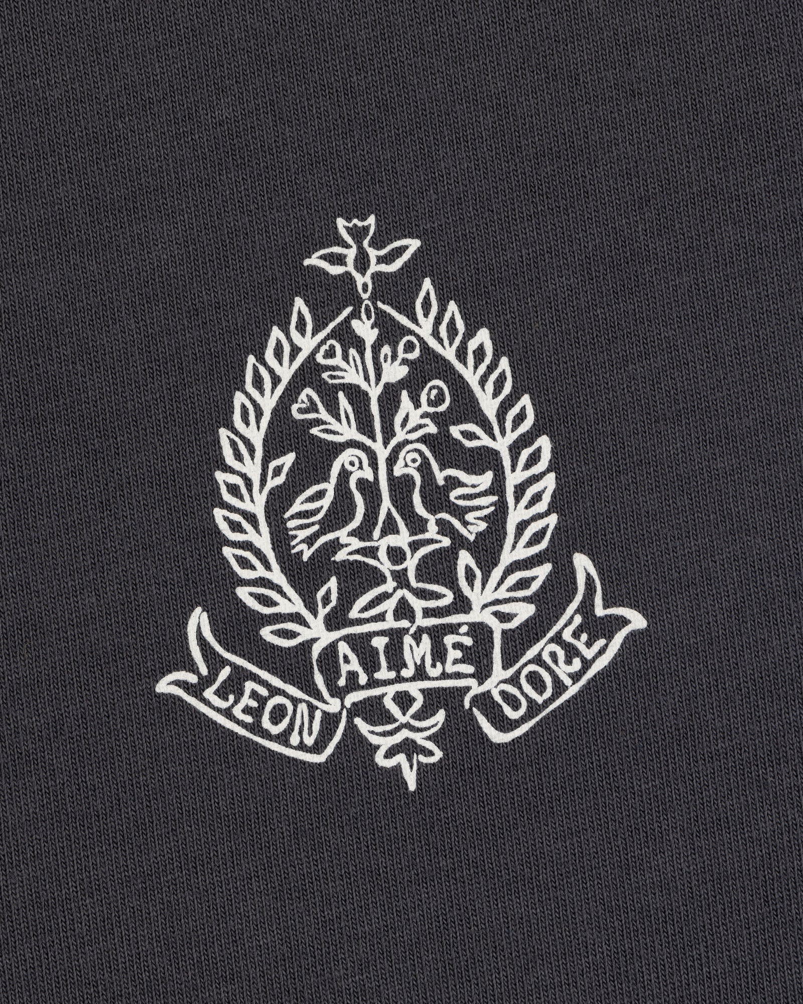 White crest with two birds, leaves, and "LEON AIME DOPE" on dark Saint George Graphic Tee.