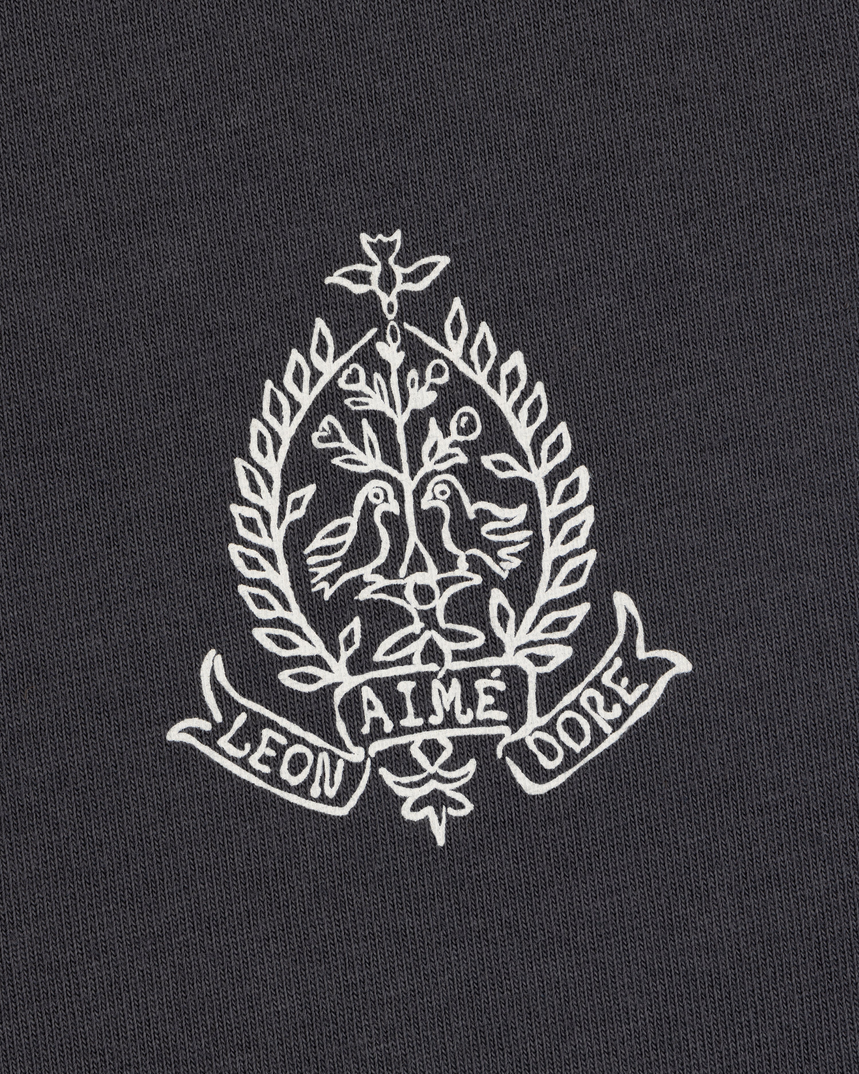 White crest with two birds, leaves, and "LEON AIME DOPE" on dark Saint George Graphic Tee.