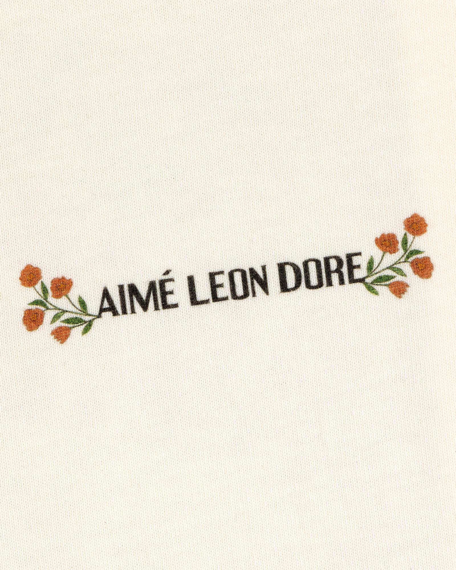 AIMÉ LEON DORE is centered with small orange flowers and green leaves on cream Aimé Postcard Tee fabric.