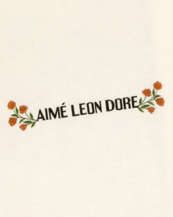 AIMÉ LEON DORE is centered with small orange flowers and green leaves on cream Aimé Postcard Tee fabric.