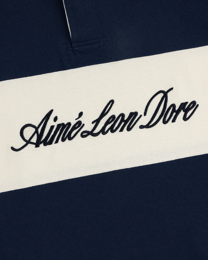 Close-up of the Script Paneled Rugby featuring Aimé Leon Dore cursive embroidery on a white horizontal stripe.