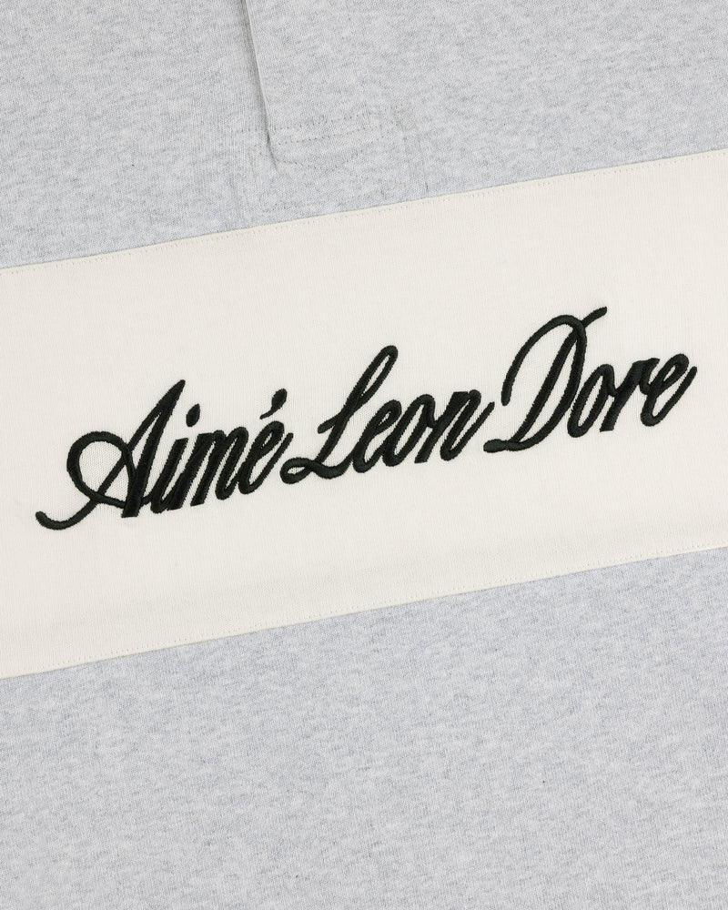 Close-up of Script Paneled Rugby with black cursive Aimé Leon Dore on a white stripe over light gray fabric.