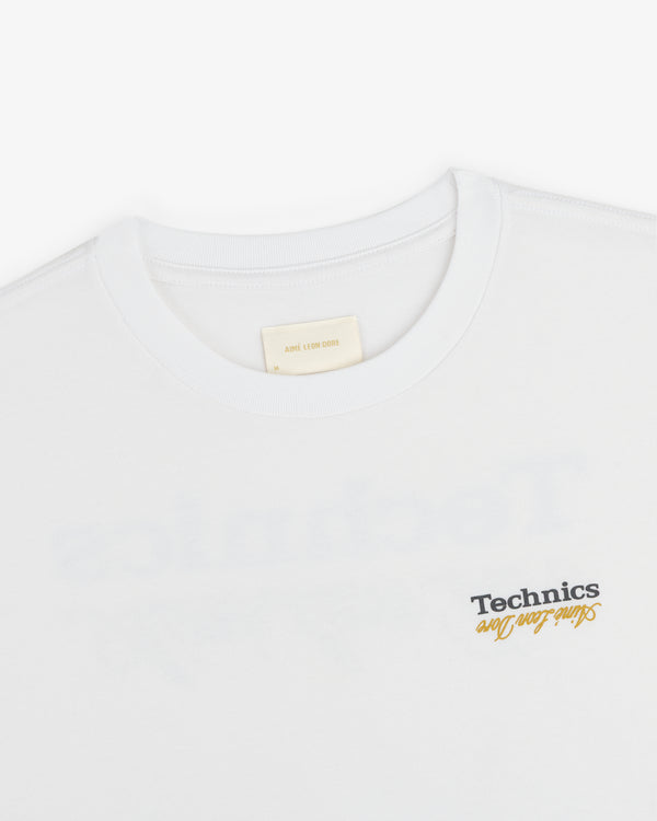 ALD / Technics Logo Tee in white with black and yellow script on front, shown from collar and upper chest.