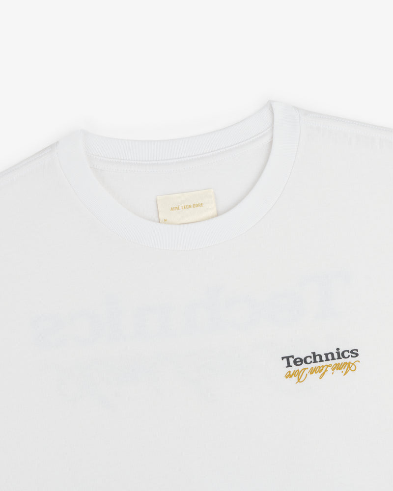 ALD / Technics Logo Tee in white with black and yellow script on front, shown from collar and upper chest.