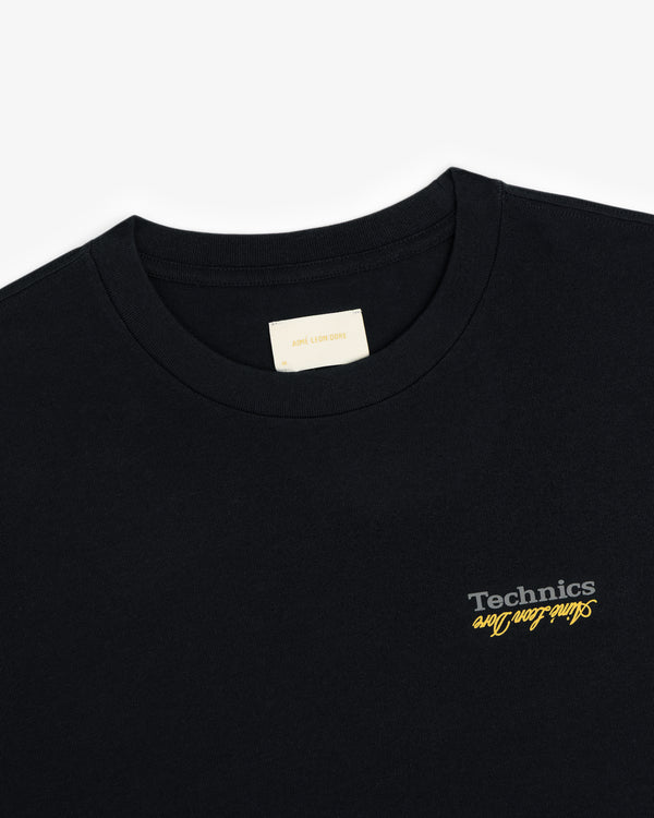 ALD / Technics Logo Tee: Black crewneck with Technics and yellow cursive text on the left chest.