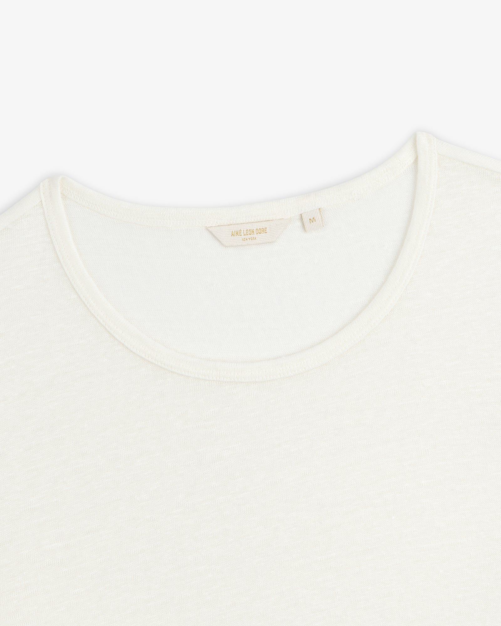 Close-up of a white Linen Tee crewneck showing its collar and inner tag with beige text on a plain background.