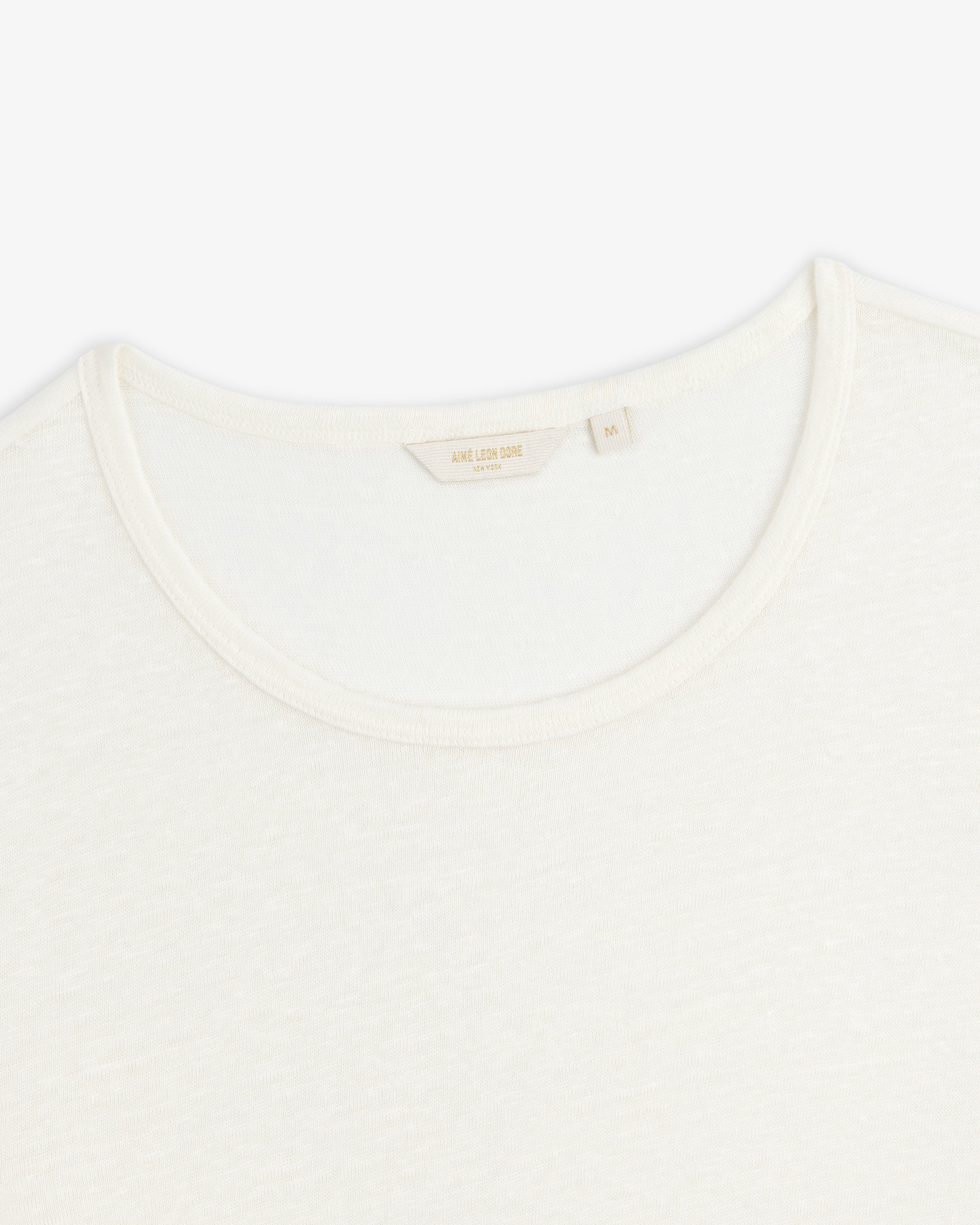 Close-up of a white Linen Tee crewneck showing its collar and inner tag with beige text on a plain background.