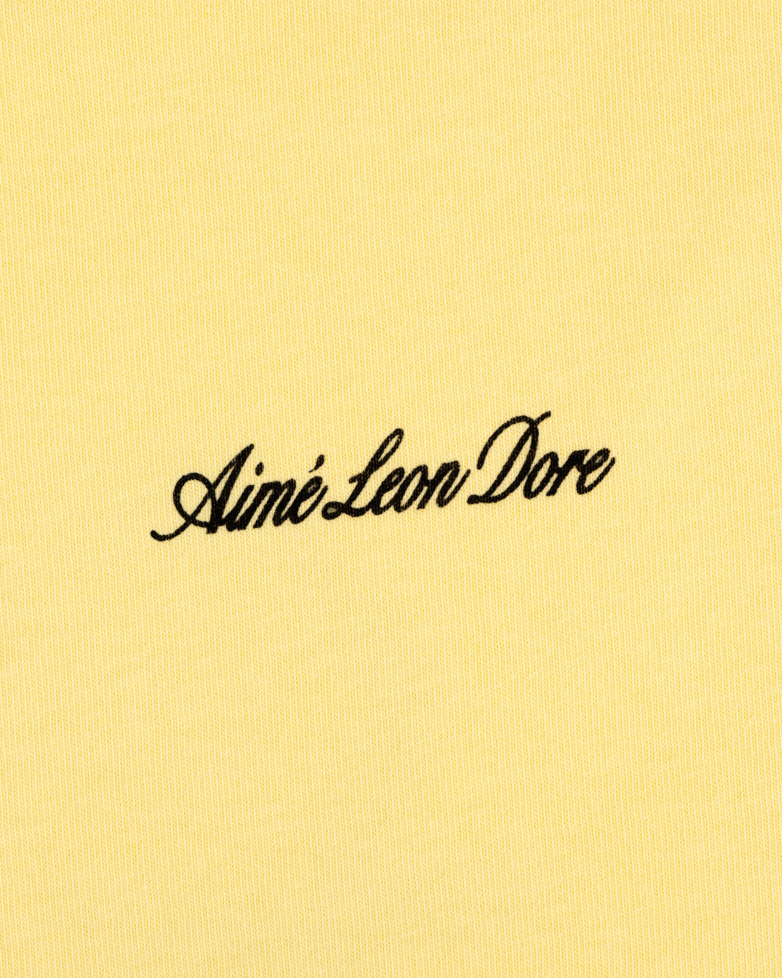 Yellow Super Cub Tee featuring Aimé Leon Dore in black cursive text at the center.