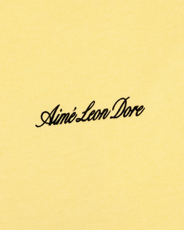 Yellow Super Cub Tee featuring Aimé Leon Dore in black cursive text at the center.
