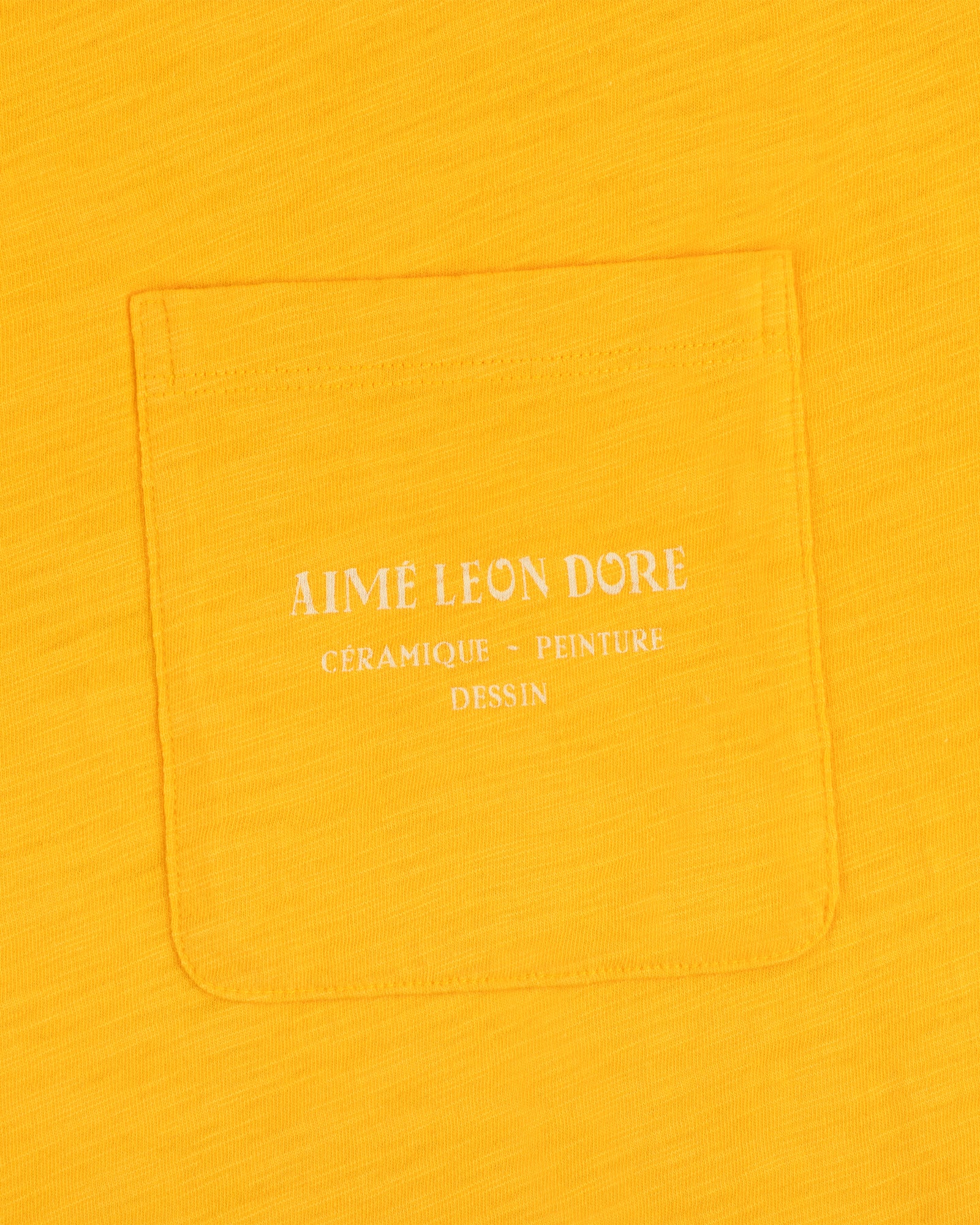 Yellow Artiste Pocket Tee with white text reading AIME LEON DORE CÉRAMIQUE - PEINTURE DESSIN.