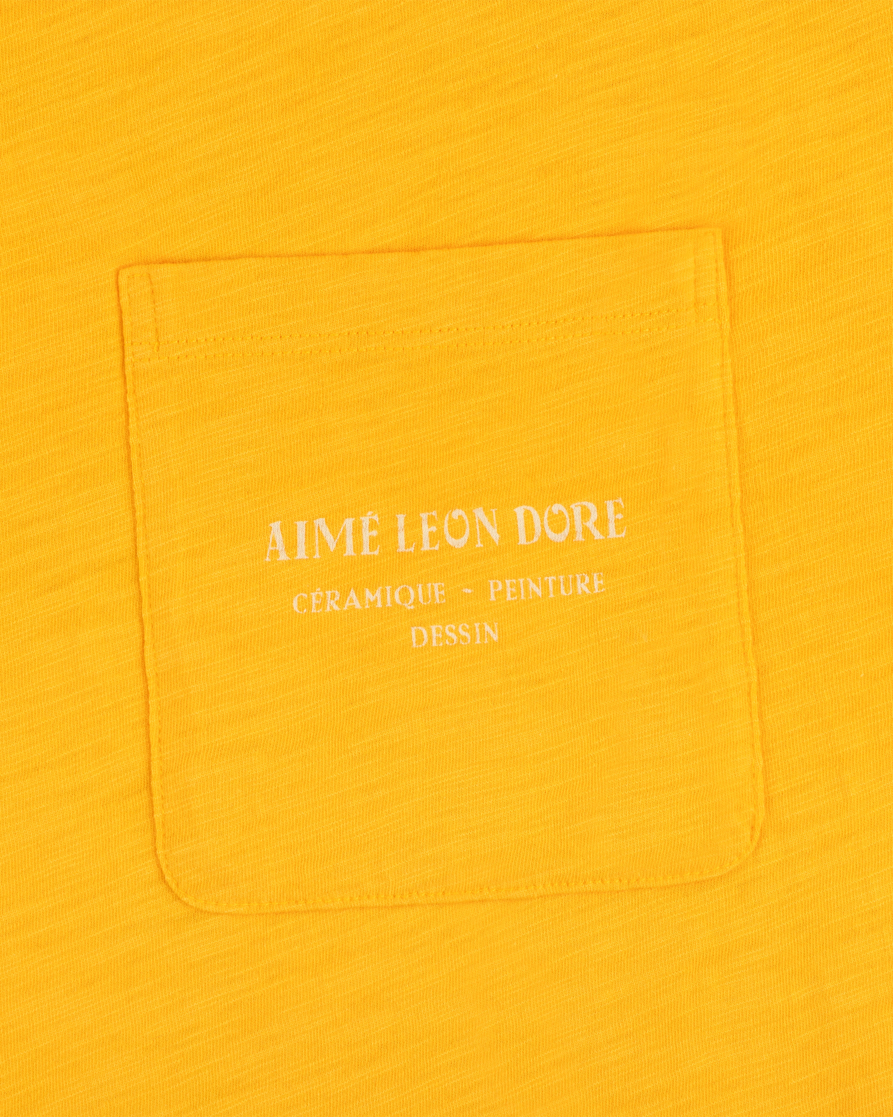 Yellow Artiste Pocket Tee with white text reading AIME LEON DORE CÉRAMIQUE - PEINTURE DESSIN.