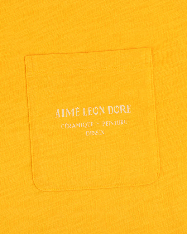 Yellow Artiste Pocket Tee with white text reading AIME LEON DORE CÉRAMIQUE - PEINTURE DESSIN.