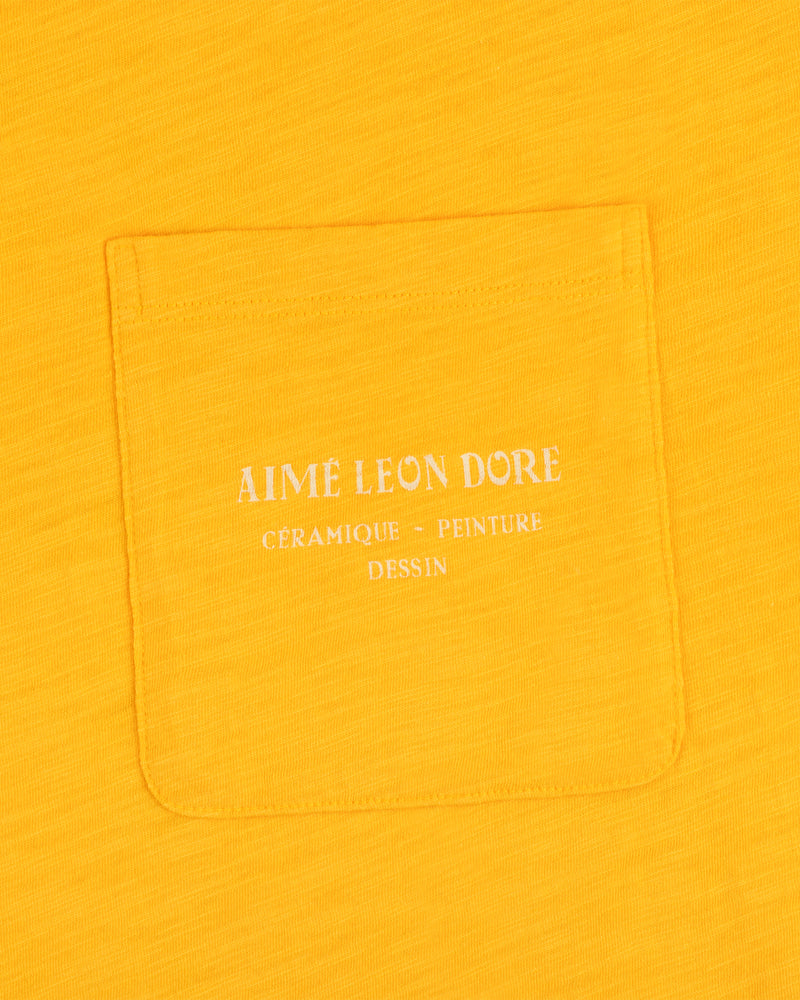 Yellow Artiste Pocket Tee with white text reading AIME LEON DORE CÉRAMIQUE - PEINTURE DESSIN.